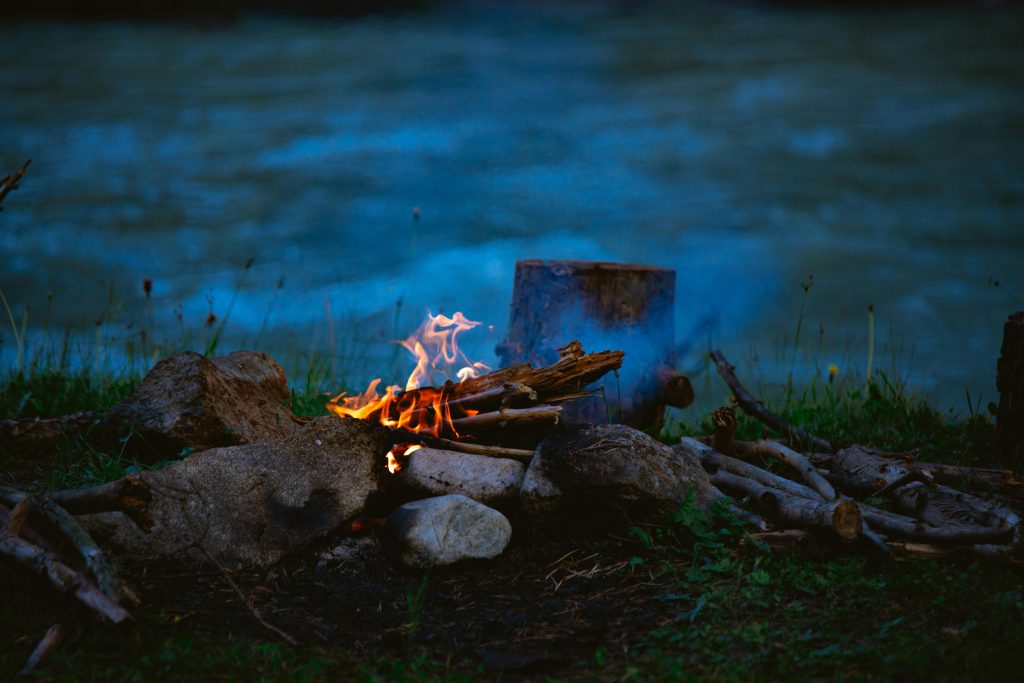 A tranquil campfire at river's edge surrounded by nature, stones, and logs.